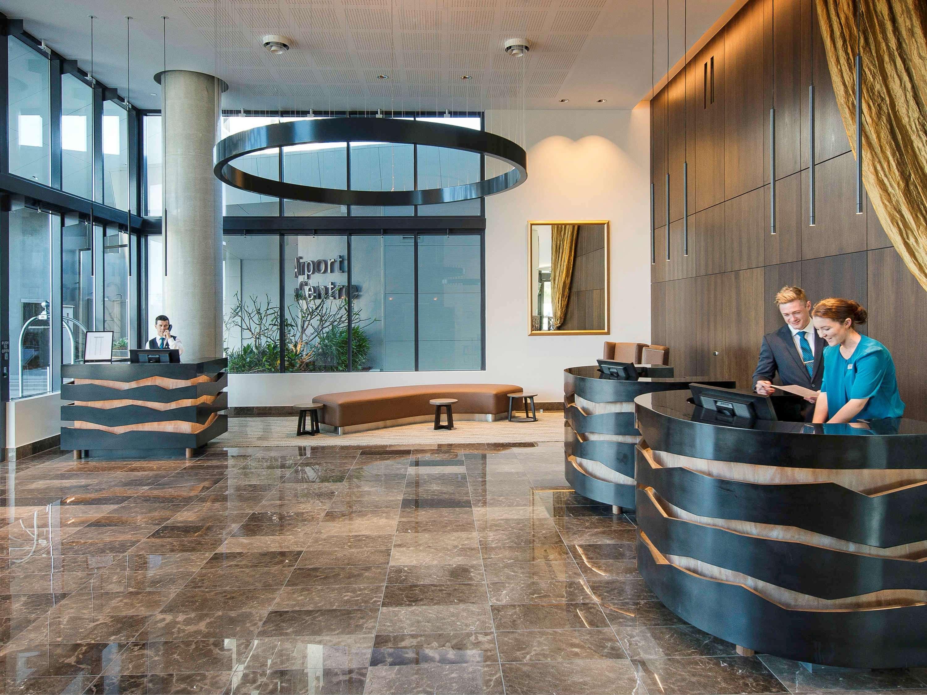 Готель Pullman Brisbane Airport Екстер'єр фото The image shows a modern hotel lobby. There are two reception desks made of dark-colored materials, and staff members are attending to guests at these desks. The lobby features large windows that allow natural light to fill the space, and there are p